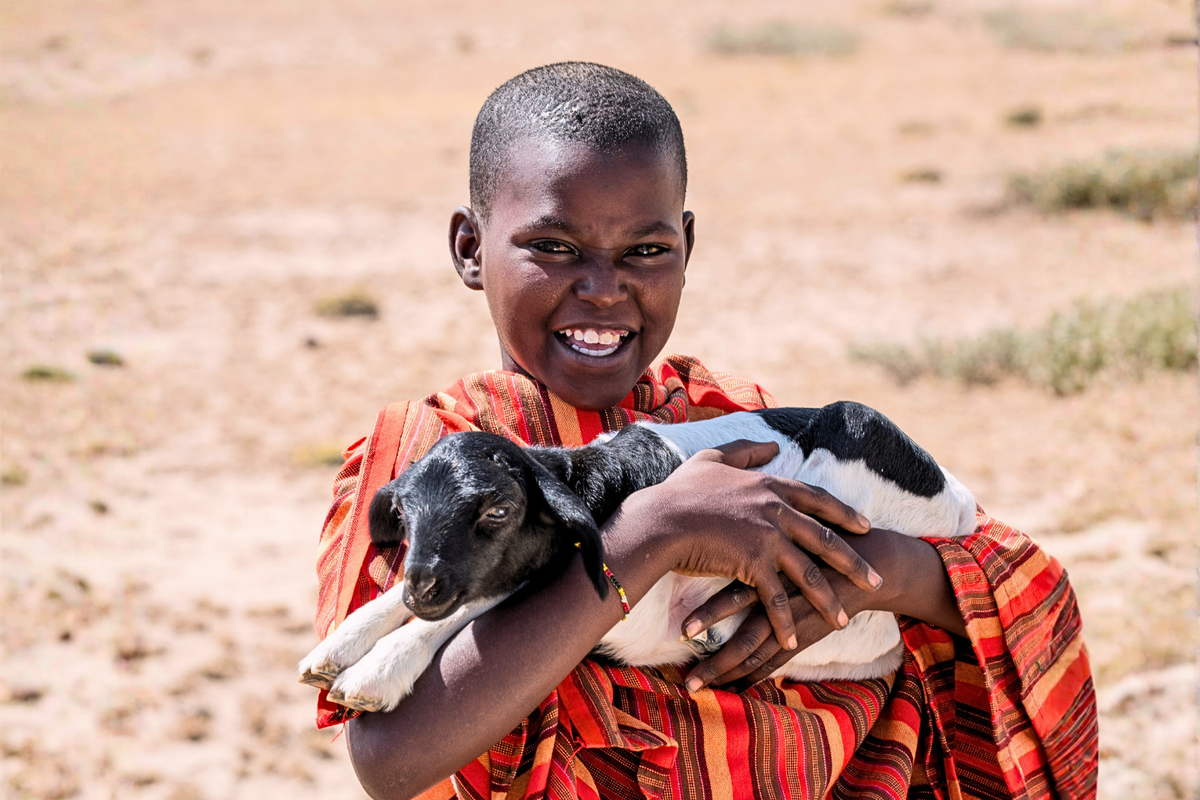A smiling young child in a red and orange striped garment holds a black-and-white goat in a dry sandy outdoor setting.