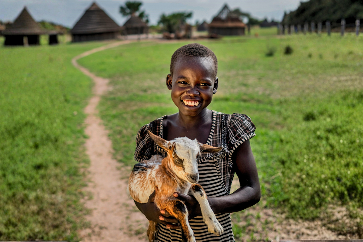 A smiling young girl in a black and white striped dress holds a small goat on a dirt path in a grassy field, with traditional huts in the background.
