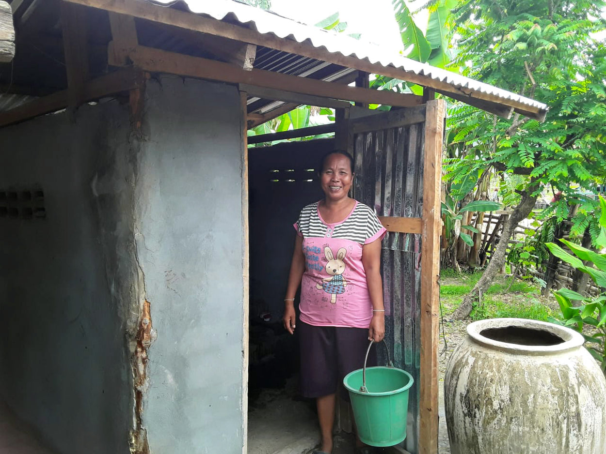 A woman stands in front a latrine with a bucket.