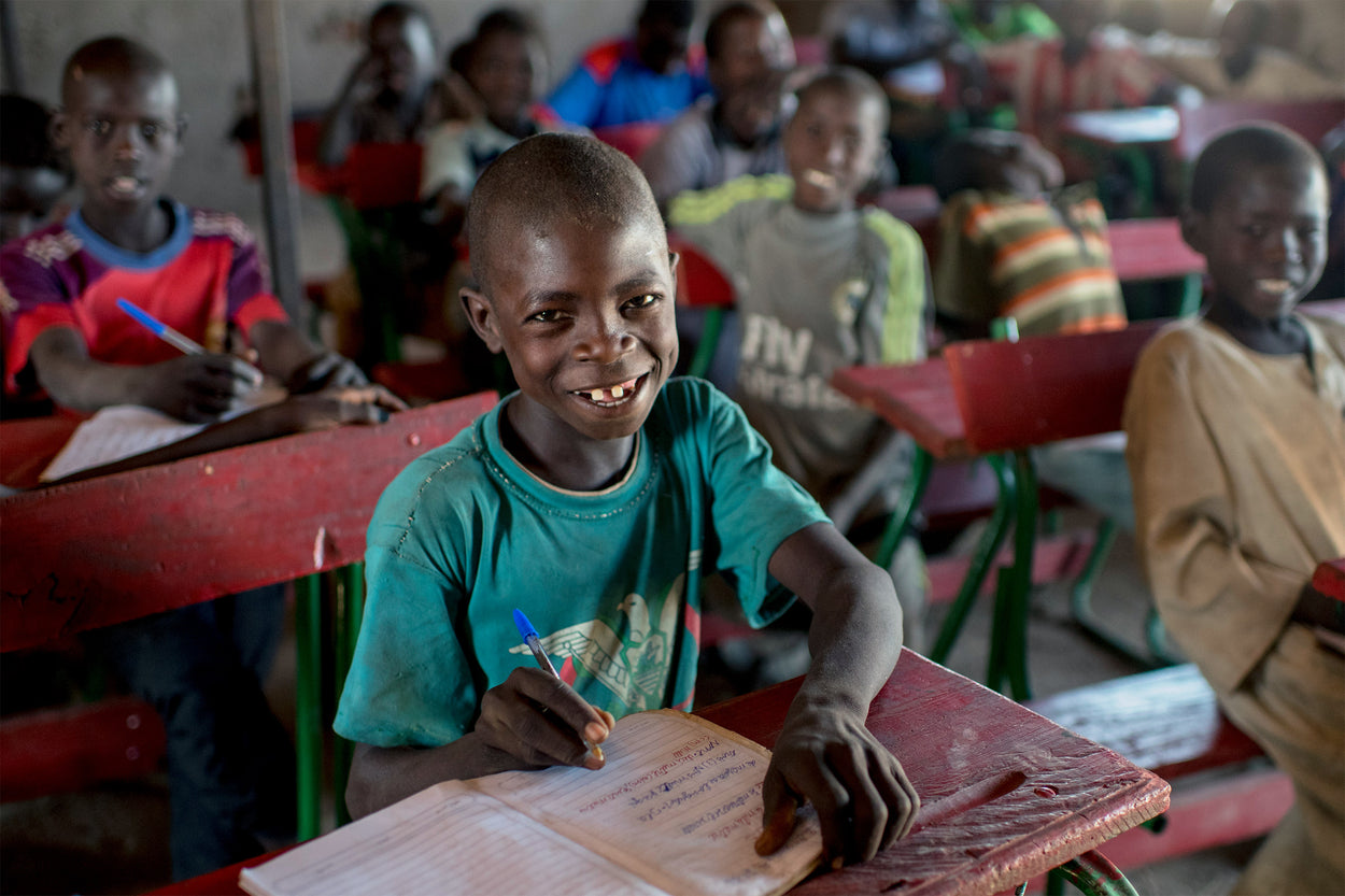 A young boy smiles at his desk because he has a classroom filled with supplies to be able to learn.